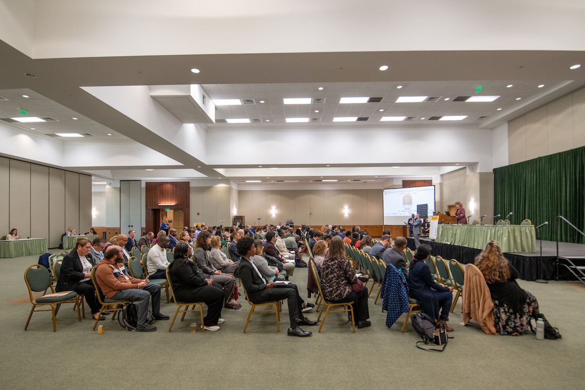 Side view of conference in session, people in several rows of chairs facing a stage.